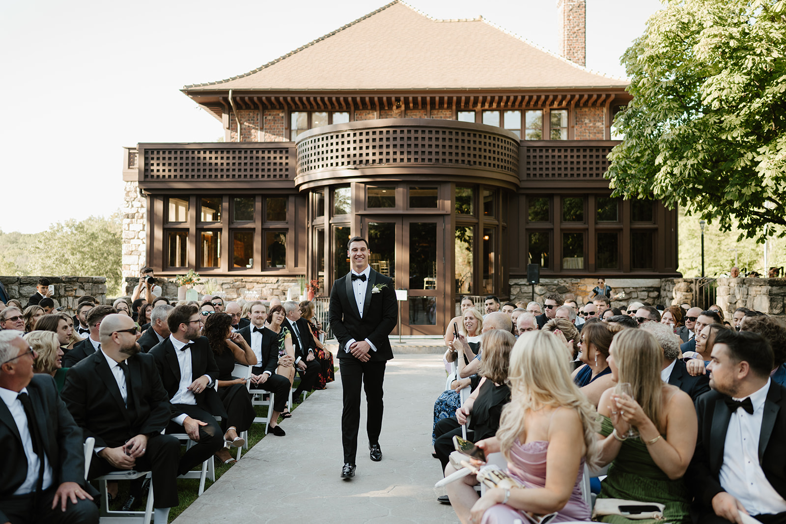 Groom walking down the aisle, smiling at seated guests with the historic mansion facade in the background.